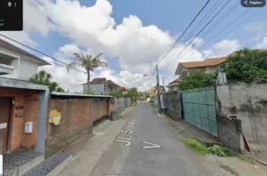 Narrow residential street with low walls and metal gates, palm trees, and overhead power lines under a partly cloudy sky.