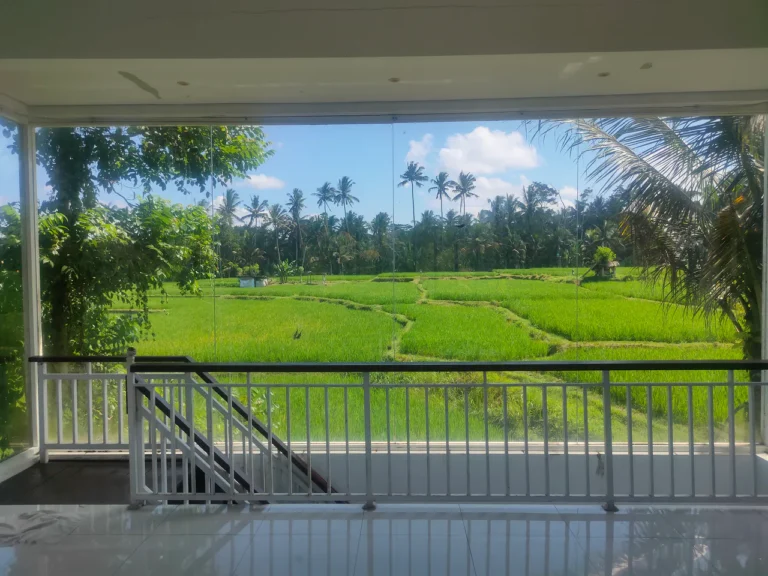 Indoor balcony with glass wall overlooking green rice fields and palm trees under a blue sky.