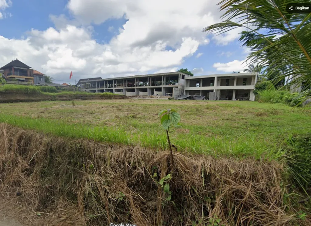 Under-construction two-story concrete building with open bays, a grassy field in front, and a small sapling in the foreground